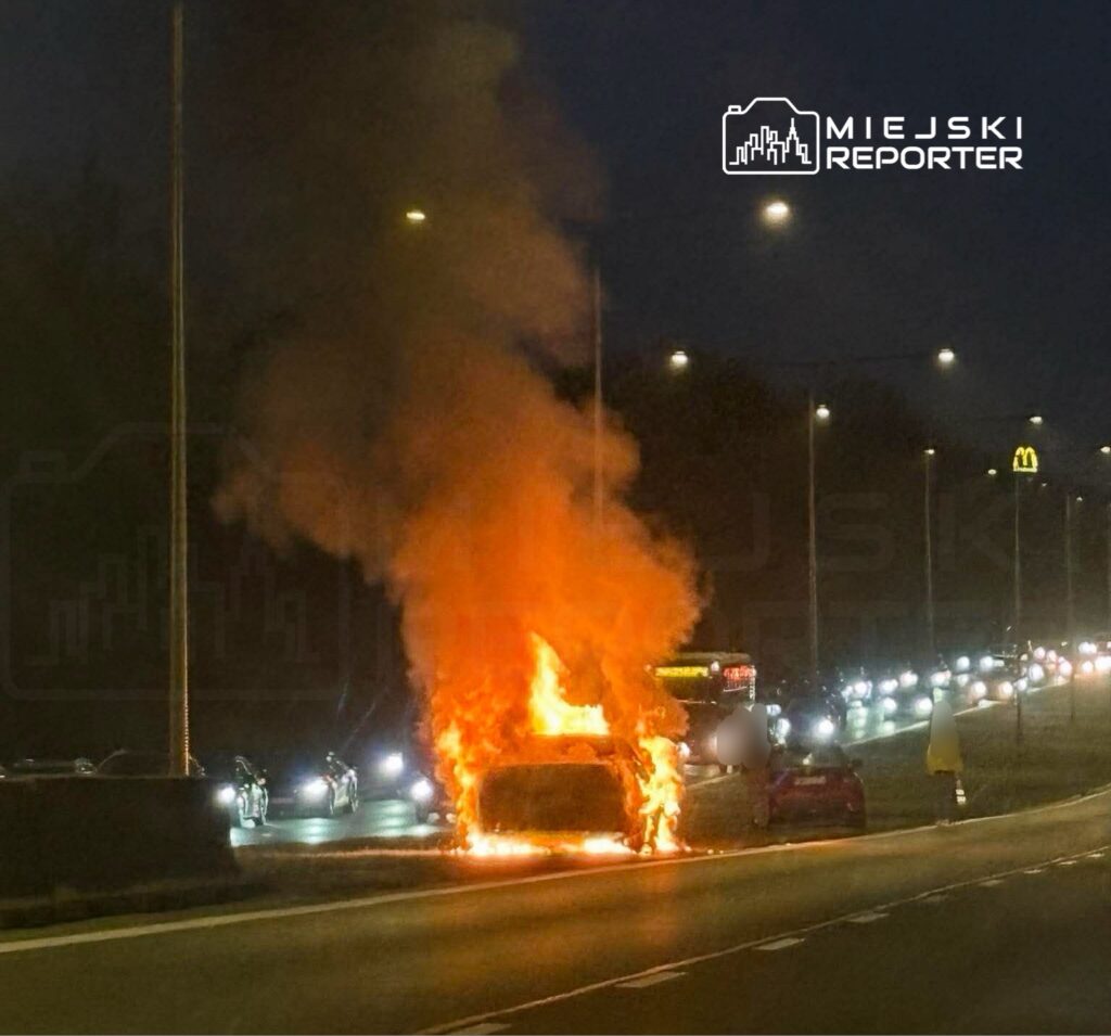 Samochód płonący na poboczu drogi, z intensywnymi płomieniami i dymem w nocy, w tle widoczne inne pojazdy.