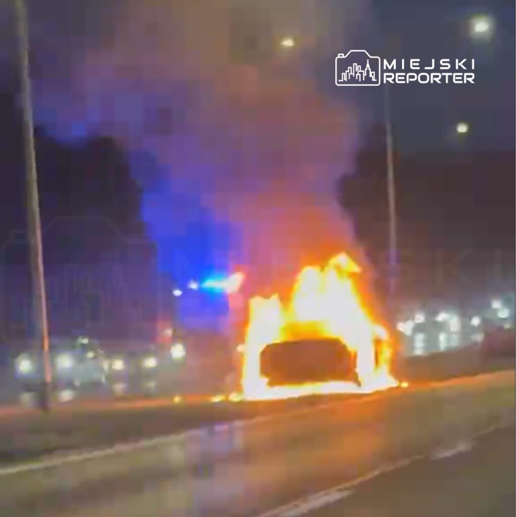 Samochód płonący na poboczu drogi, otoczony przez policyjne światła w nocy.
