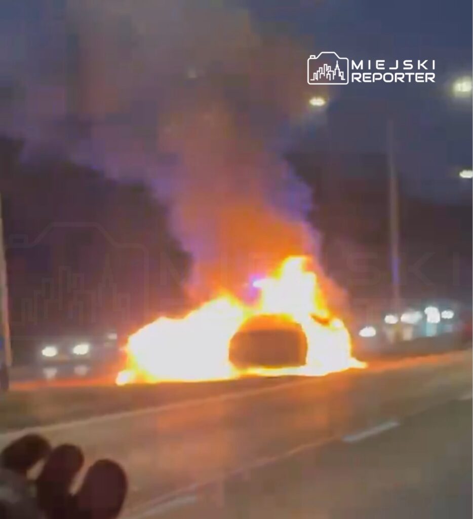 Samochód w płomieniach na poboczu drogi, otoczony dymem, w tle widoczne światła innych pojazdów.