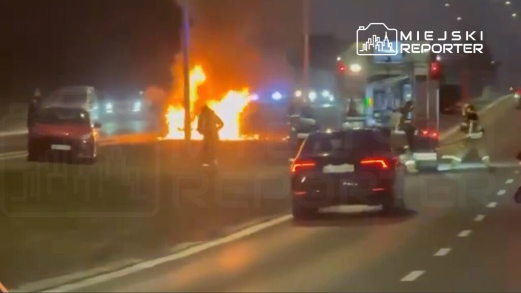 Pojazd płonący na poboczu drogi, w tle widać pojazdy ratunkowe i osoby próbujące ugasić ogień.