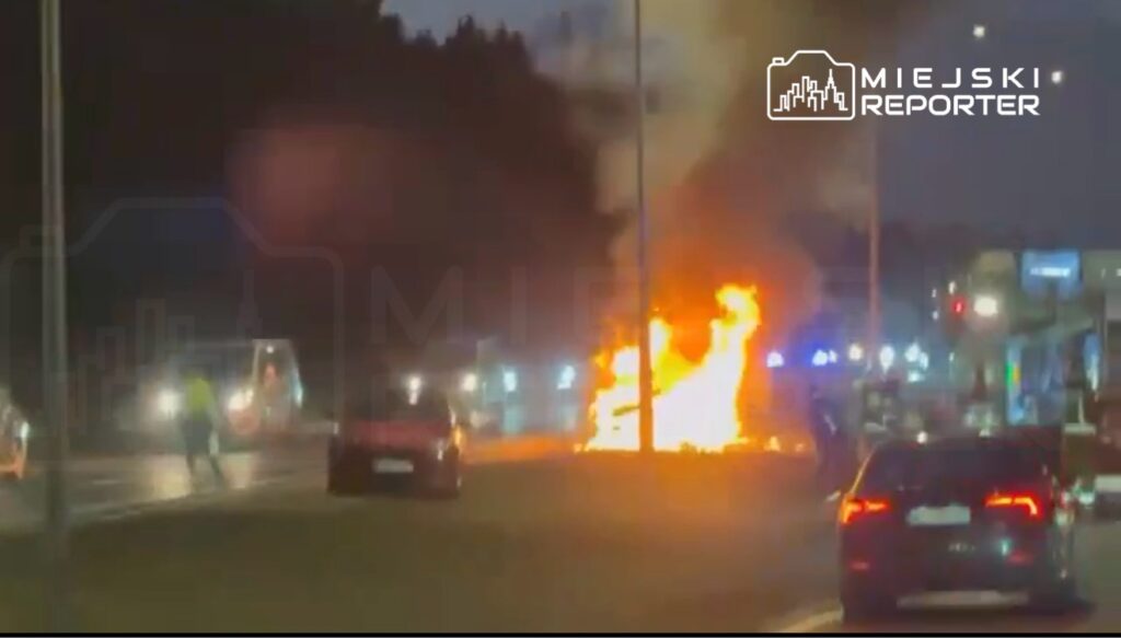 Pojazd płonący na poboczu drogi, obok interweniujące służby i przejeżdżające samochody.