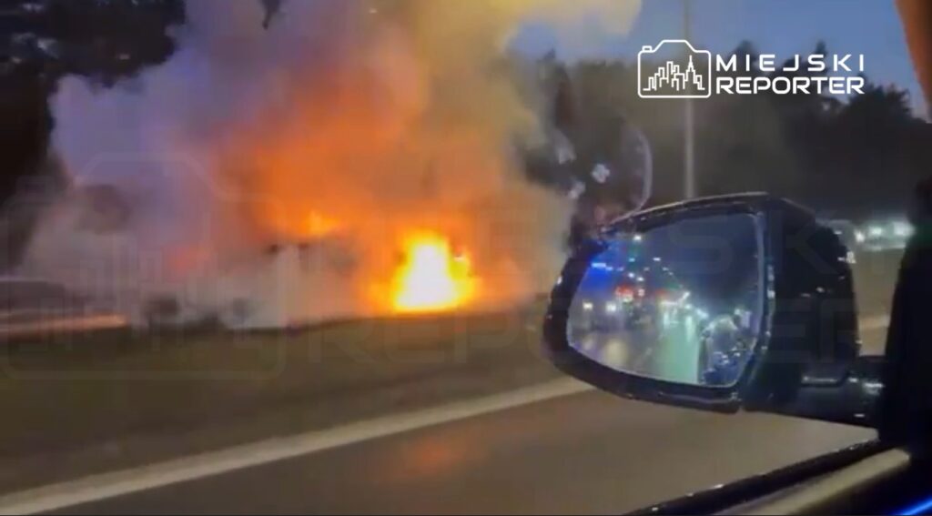 Pożar trawiący krzewy przy drodze, widoczny z wnętrza samochodu, w tle widać inne pojazdy.