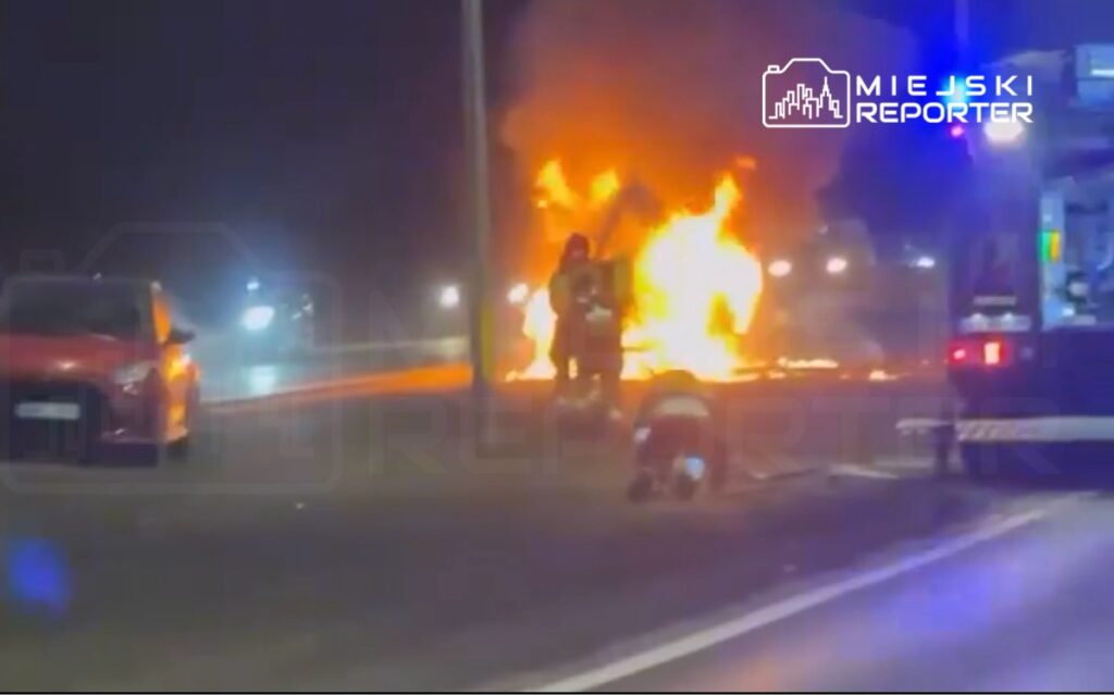 Strażacy gaszą płonący samochód na drodze w nocy, w tle widoczne inne pojazdy.