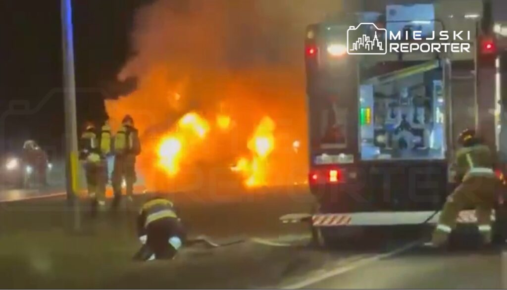 Strażacy gaszą płonący pojazd na drodze, podczas gdy jeden z nich podłącza wąż gaśniczy do wozu strażackiego.
