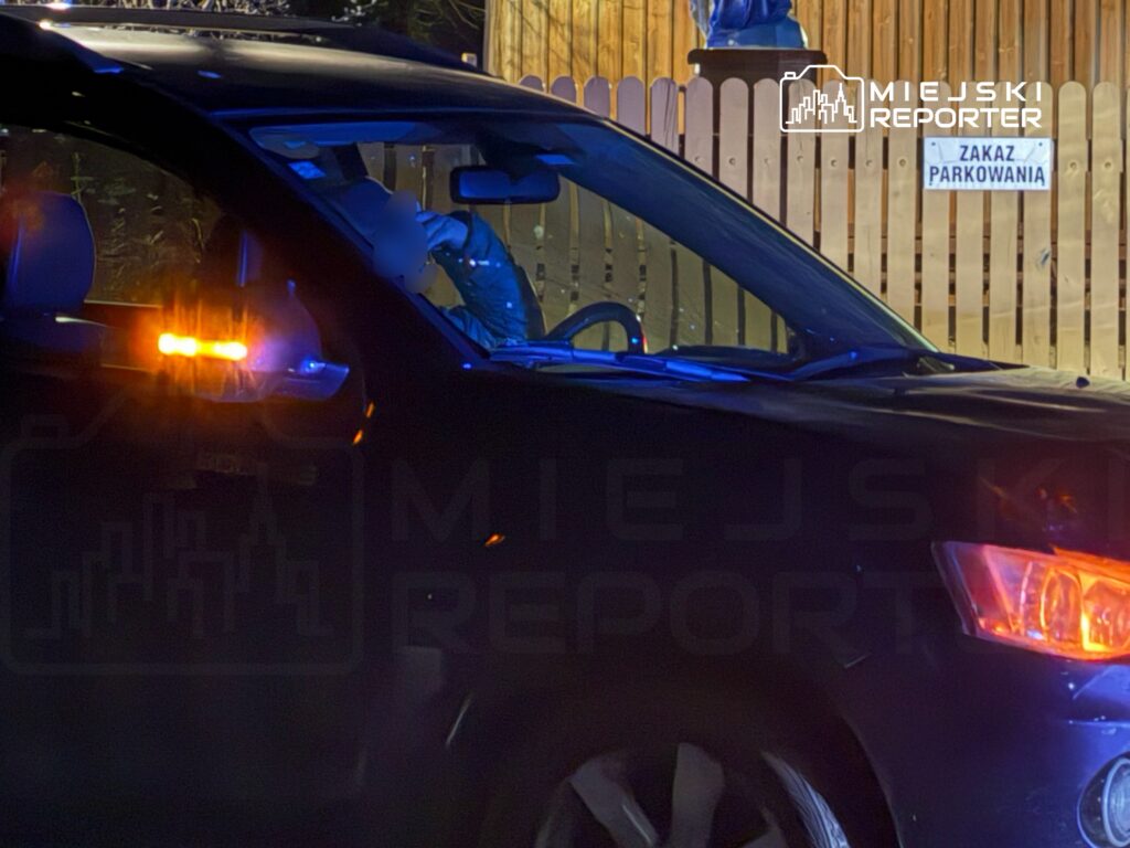 Samochód osobowy z włączonymi światłami awaryjnymi stoi obok znaku „Zakaz parkowania” w oświetlonym otoczeniu.