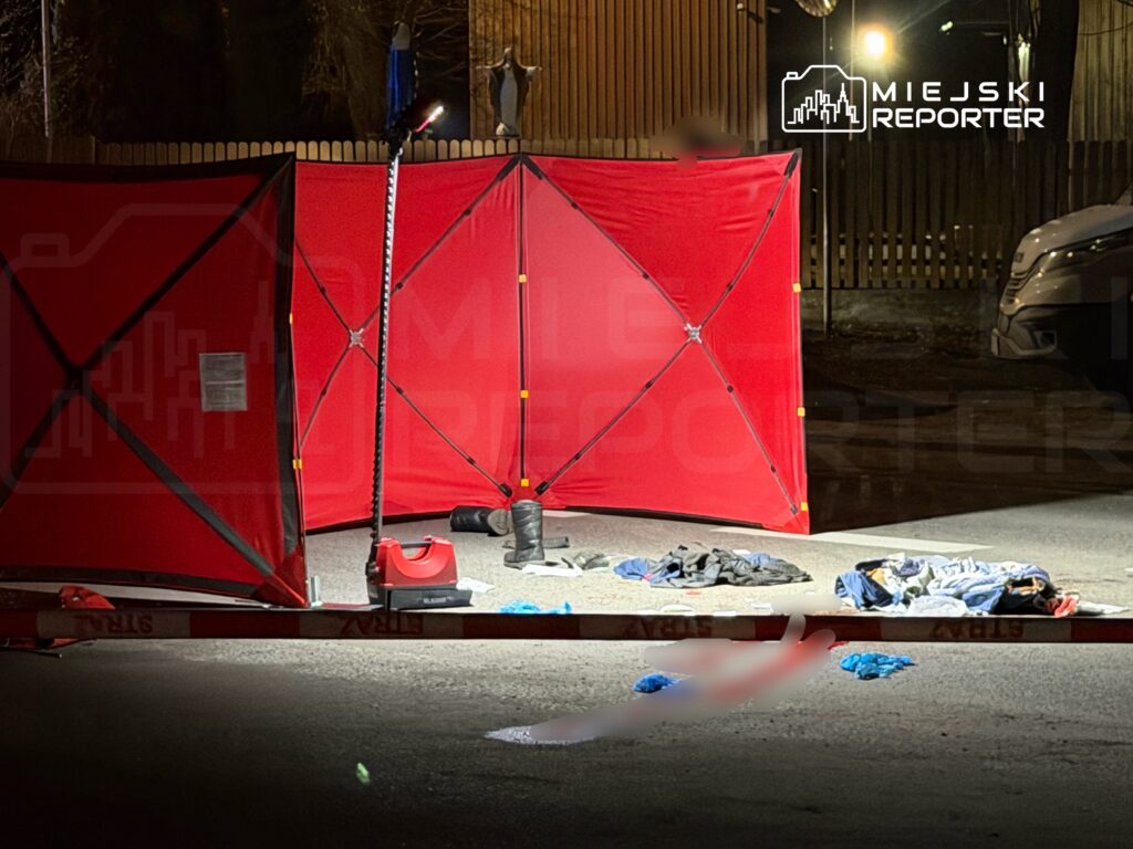 Czerwony namiot policyjny ustawiony na drodze, obok leżące ubrania oraz przedmioty osobiste. W tle oświetlone otoczenie.