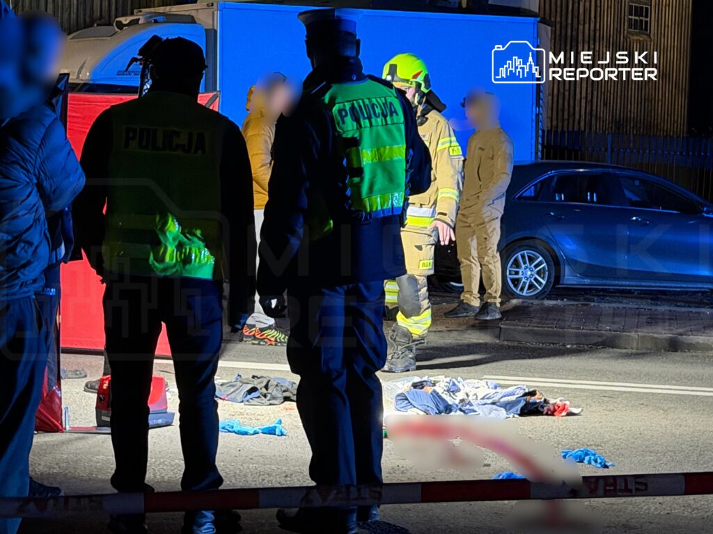 Grupa policjantów i strażaków pracuje na miejscu zdarzenia drogowego, obok leżących ubrań na jezdni.