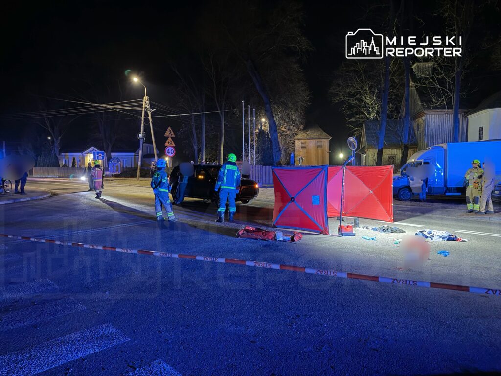 Ratownicy i policja pracują na miejscu wypadku drogowym, otoczeni przez czerwone parawany i oznakowanie.