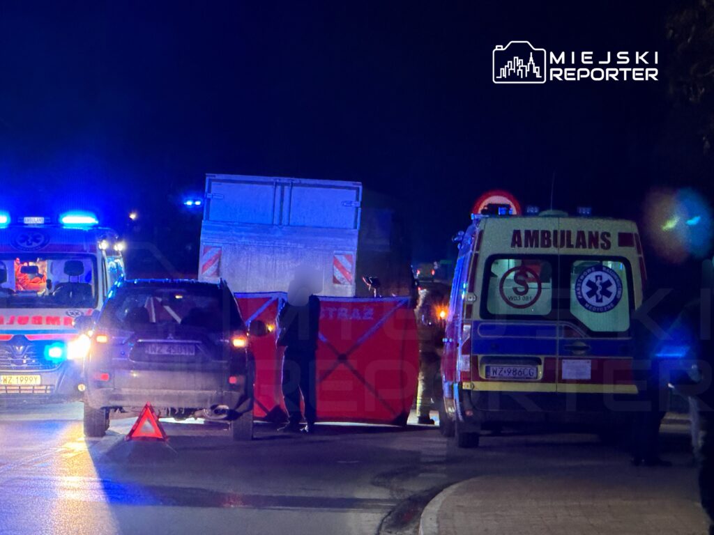 Na drodze nocą zablokowane pojazdy, w tym karetka i samochód osobowy, a obok stoi mężczyzna z trójkątnym znakiem ostrzegawczym.
