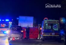 Na drodze nocą zablokowane pojazdy, w tym karetka i samochód osobowy, a obok stoi mężczyzna z trójkątnym znakiem ostrzegawczym.