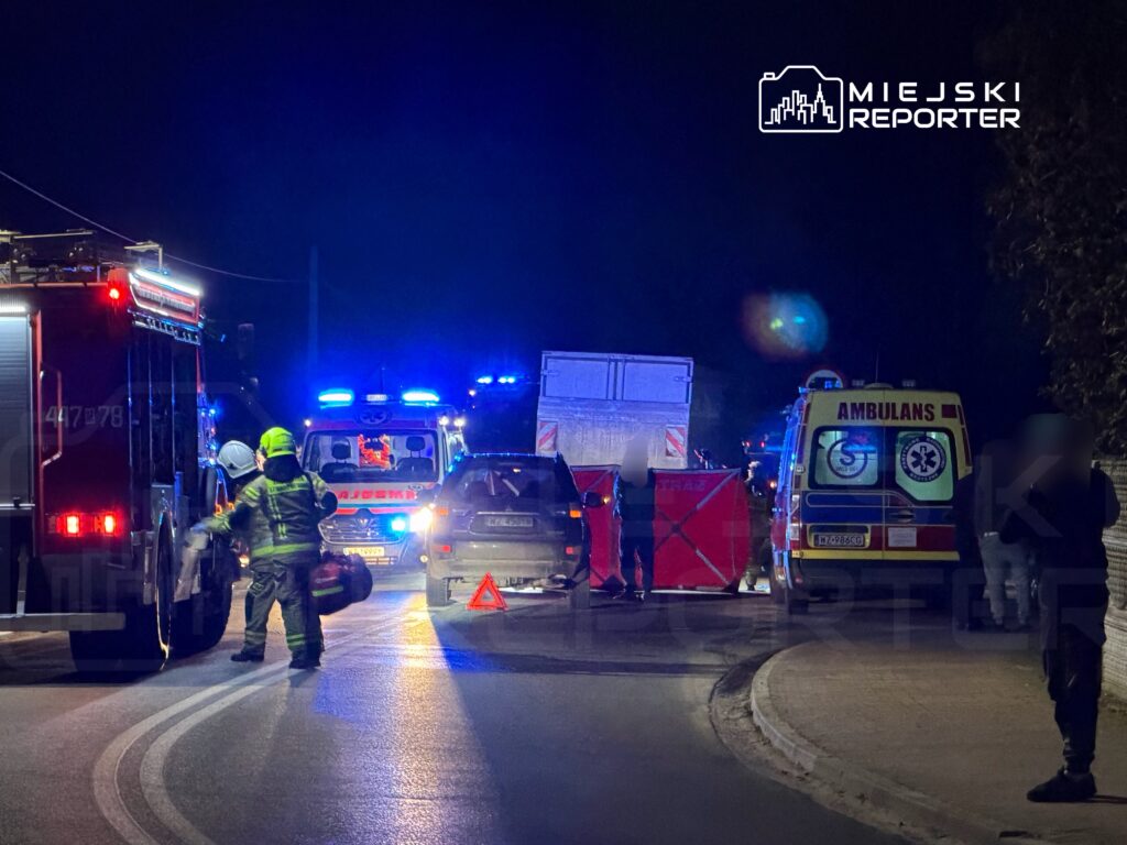 Strażak w hełmie przygotowujący sprzęt obok wozu strażackiego na drodze, w tle pojazdy służb ratunkowych.