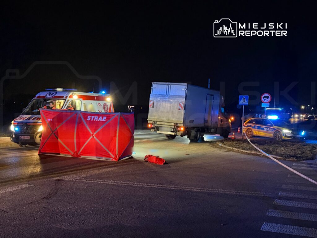 Karetka pogotowia oraz radiowóz policji zabezpieczają miejsce wypadku drogowego w nocy, z czerwonym parawanem w tle.