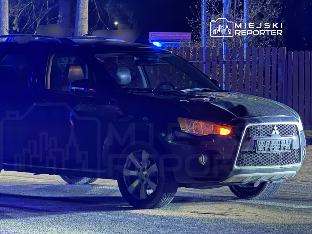 Czarny SUV zaparkowany na ulicy, oświetlony niebieskimi światłami. W tle widoczny drewniany płot i znaki drogowe.