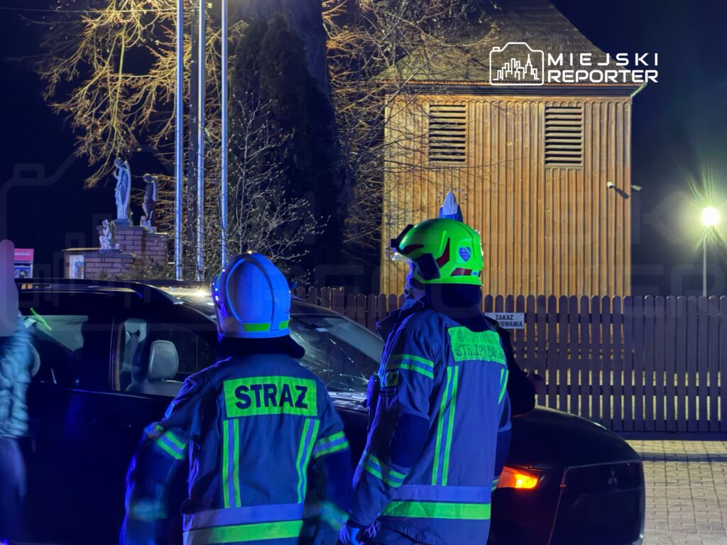 Dwaj strażacy w uniformach z odblaskowymi elementami obserwują sytuację przy ciemnym samochodzie w nocy. W tle widoczny drewniany budynek.