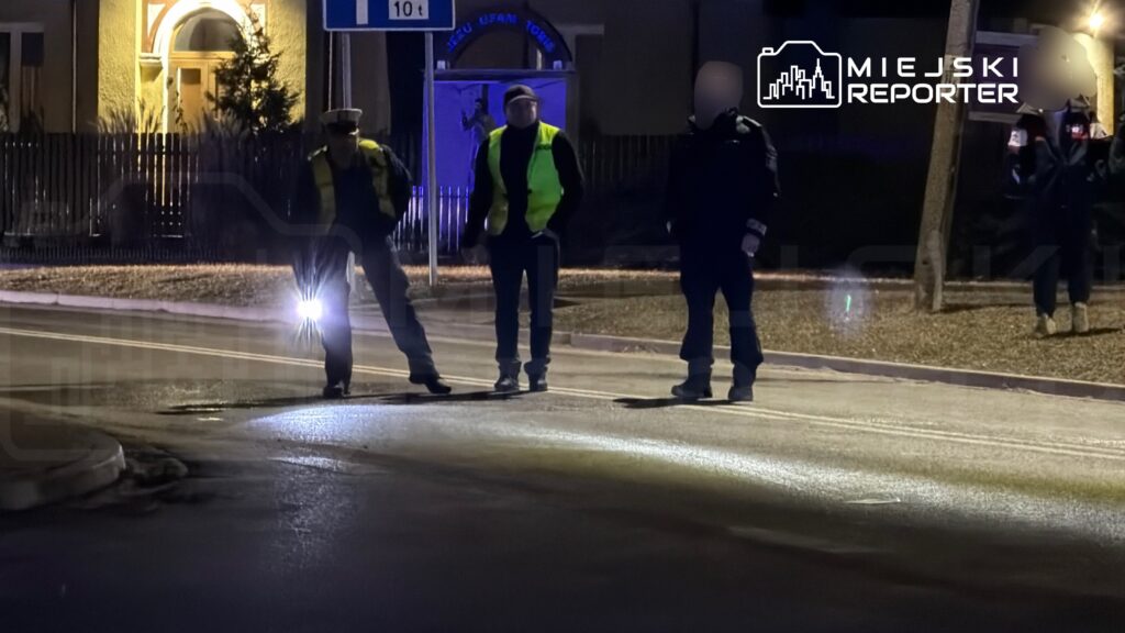 Trzech funkcjonariuszy w odblaskowych kamizelkach prowadzi czynności na oświetlonej ulicy nocą.