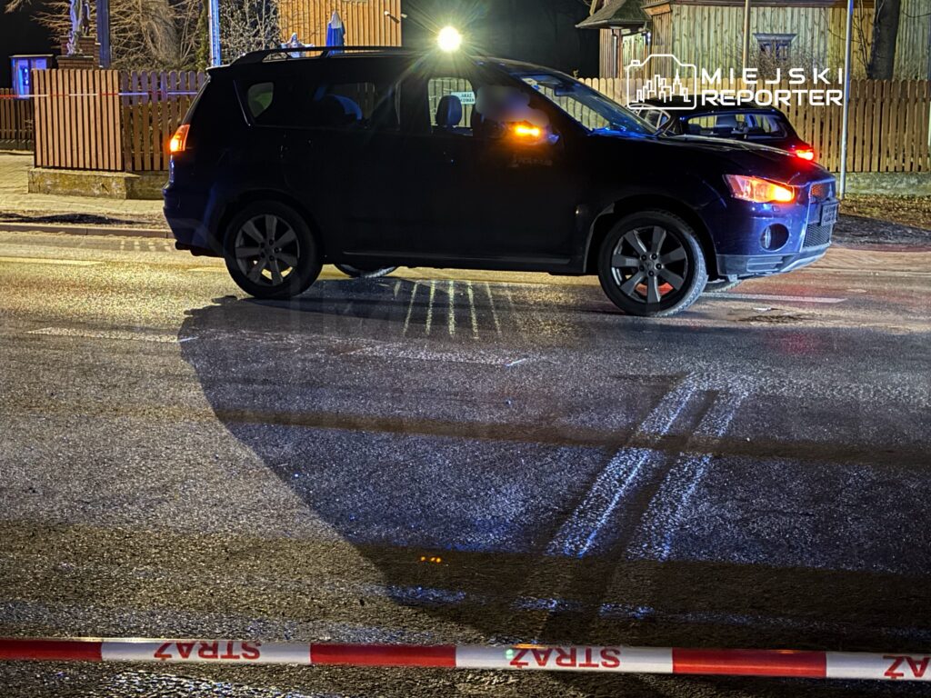 Czarny SUV zatrzymany na oświetlonej ulicy, na tle drewnianego ogrodzenia i figurki świętej. Widać barierki z napisem "STREFA".