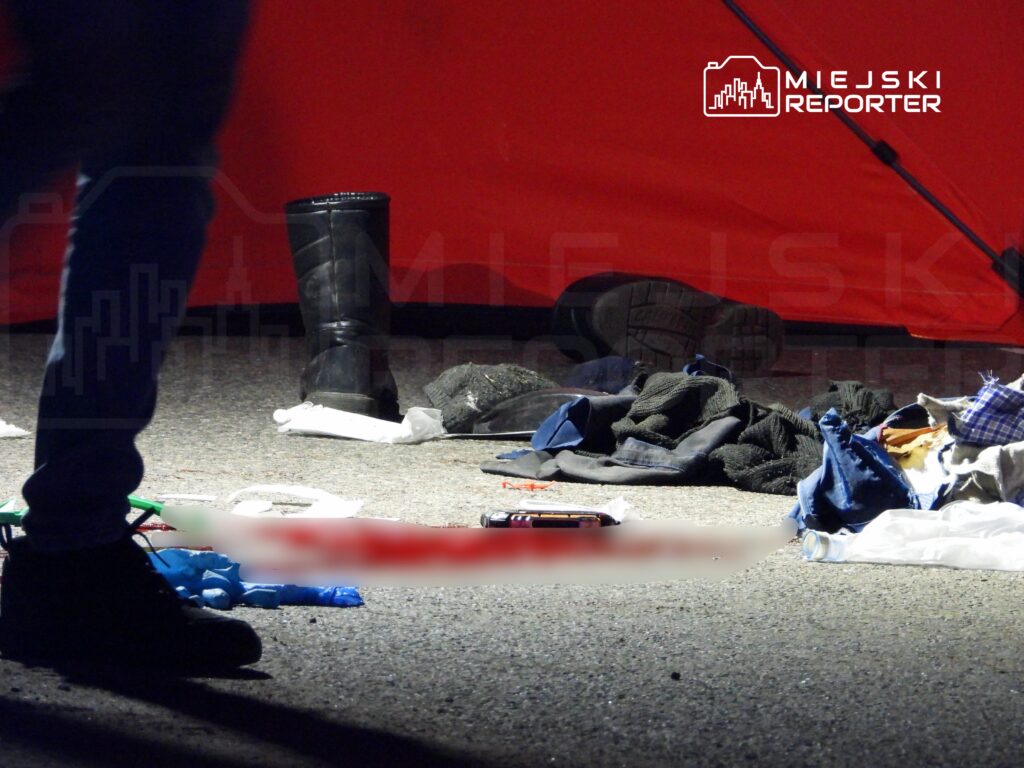 Na ulicy leżą porozrzucane ubrania i przedmioty, a w tle widać czerwoną namiotową strukturę.