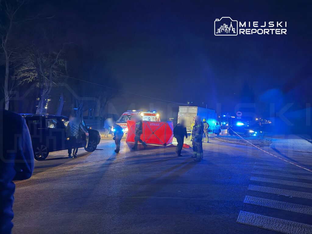 Grupa ratowników i policjantów pracuje przy miejscu zdarzenia drogowego, w tle widać ambulans i radiowóz.