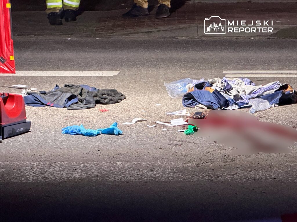 Na ulicy leżą porozrzucane ubrania oraz przedmioty, a w tle widoczni są strażacy w odzieży roboczej.