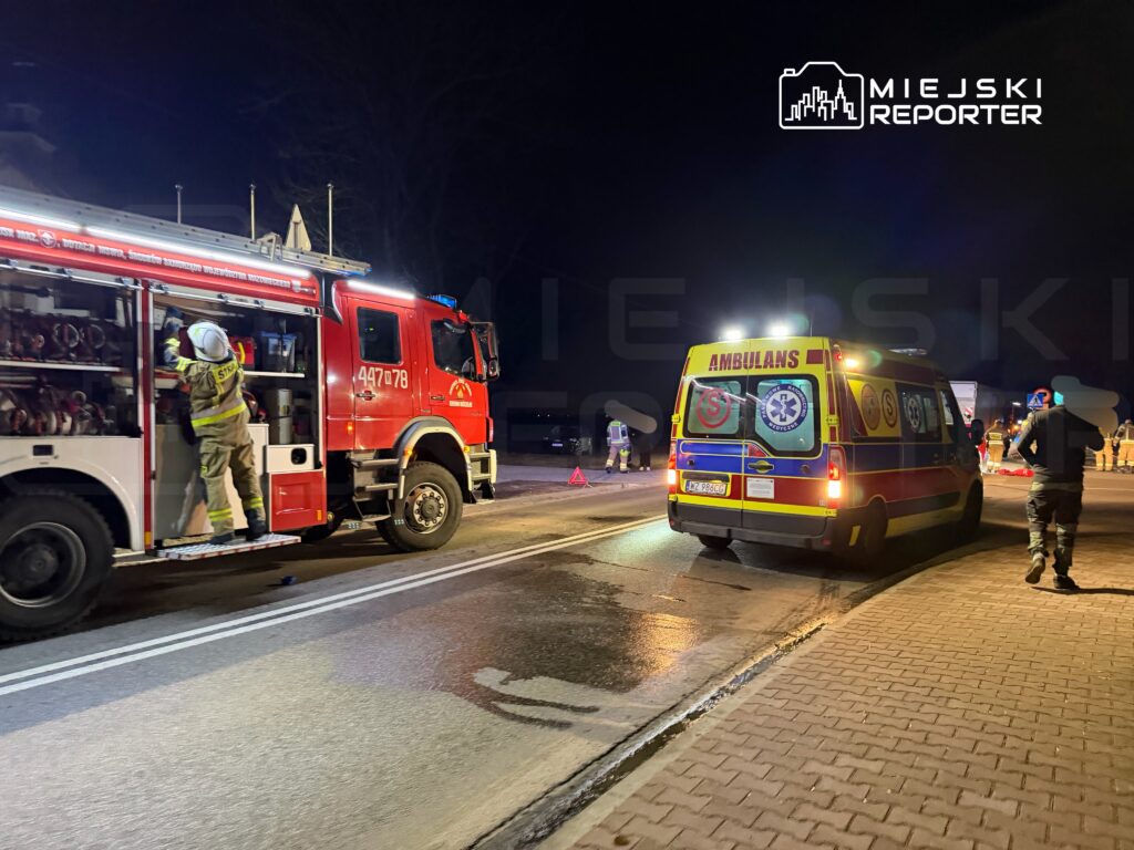 Strażak w kasku wyciąga sprzęt z wozu strażackiego, obok stoi ambulans na oświetlonej drodze nocą.