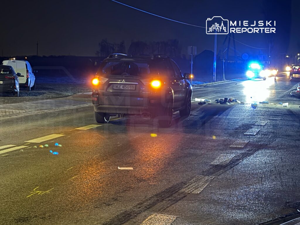 Samochód osobowy zatrzymany na drodze, obok leżące przedmioty, w tle niebieskie światła radiowozu policyjnego.