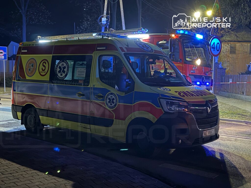 Ambulans z włączonymi światłami sygnalizacyjnymi stoi na ulicy nocą, obok wozu strażackiego.