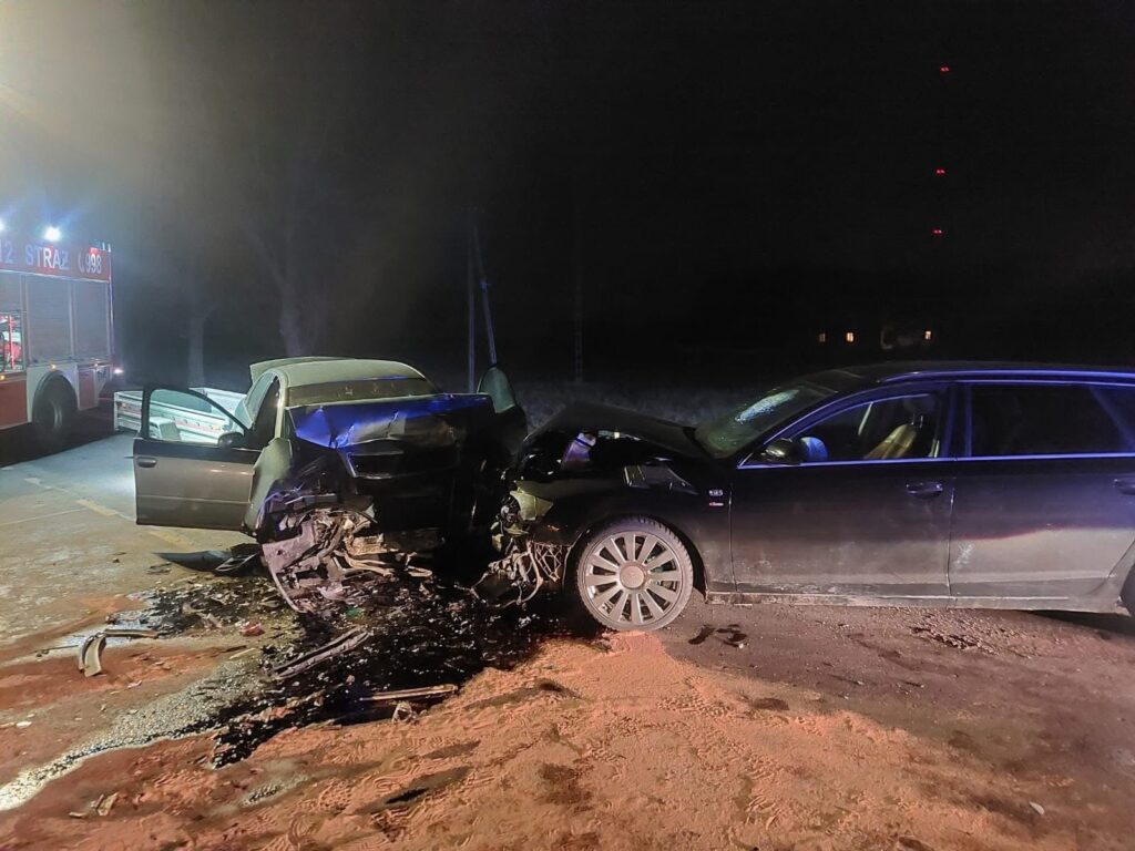 Dwa uszkodzone samochody osobowe zderzone na drodze w nocy, obok widoczny wóz strażacki.