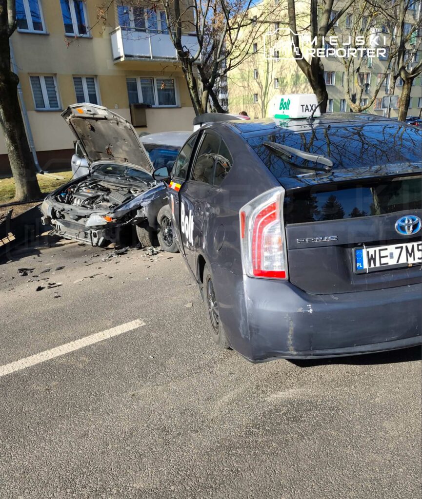 Zniszczony samochód osobowy z otwartą maską stoi na ulicy obok taksówki Bolt, w tle widać blok mieszkalny.