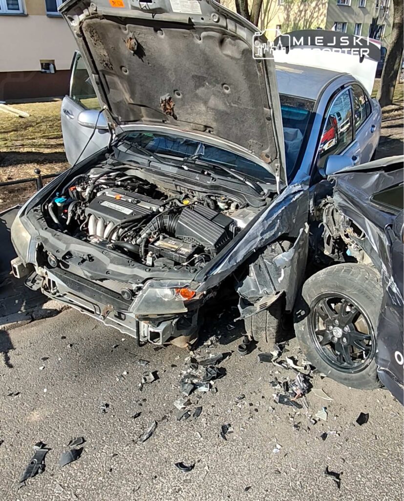 Uszkodzony samochód z otwartą maską stoi na drodze, obok widać fragmenty zderzaków i innych elementów.