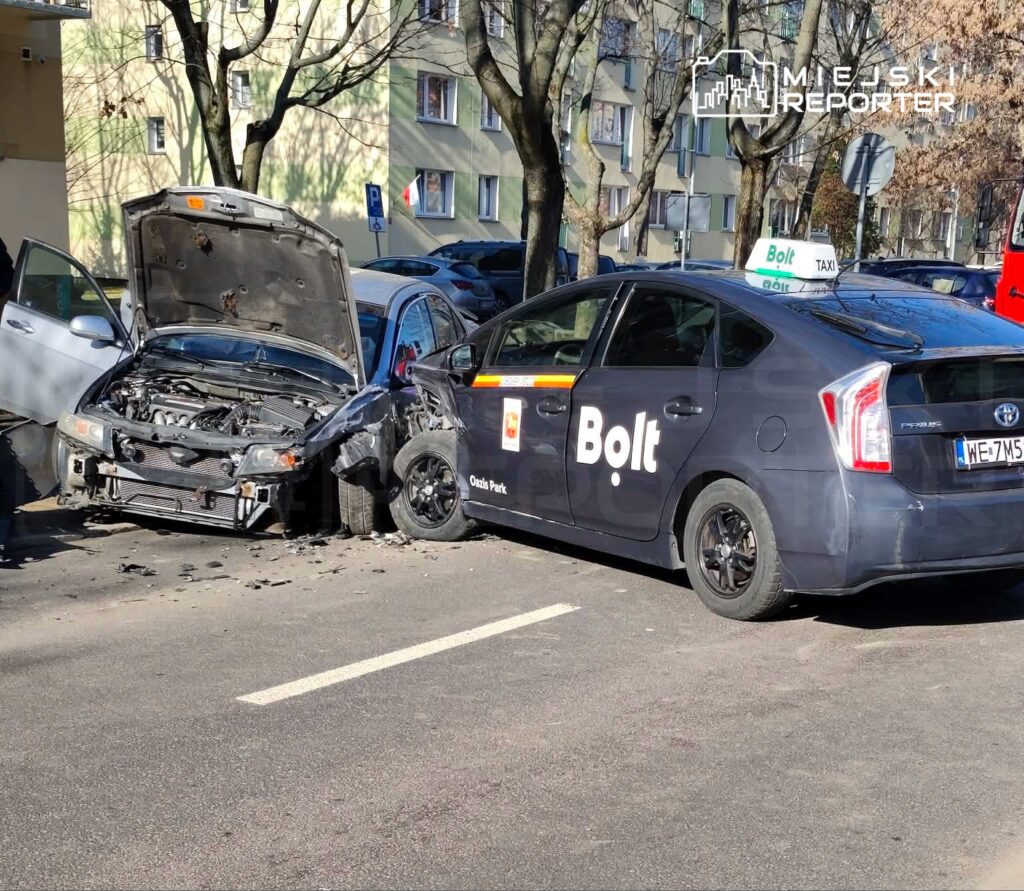 Zderzenie dwóch samochodów na parkingu, z uszkodzoną maską jednego z pojazdów i oznaczeniem "Bolt" na taksówce.
