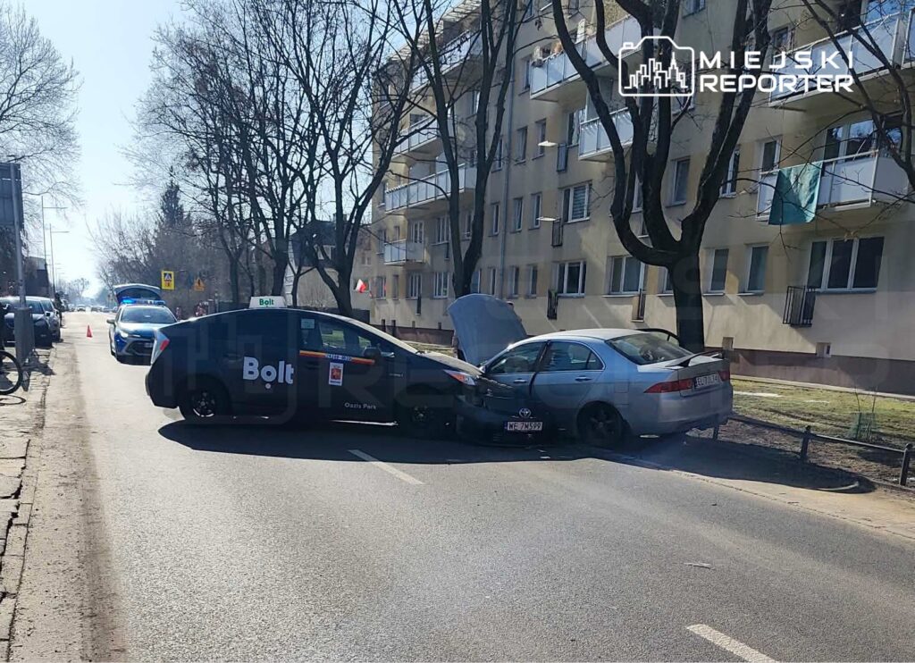 Czarny samochód osobowy z logo Bolt zderzył się z szarym pojazdem na ulicy, obok bloków mieszkalnych.