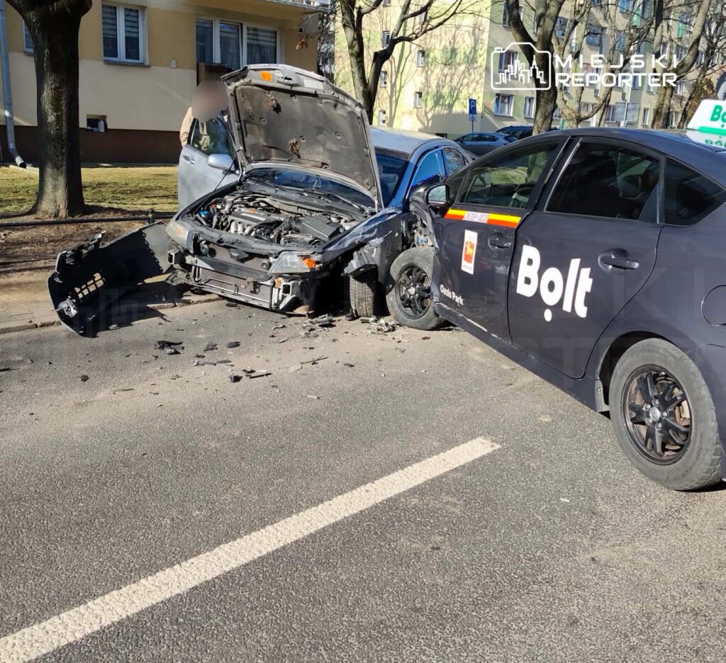 Zderzenie dwóch samochodów na ulicy, jeden z pojazdów z uszkodzoną maską i widocznymi odłamkami, obok stoi auto z logo Bolt.