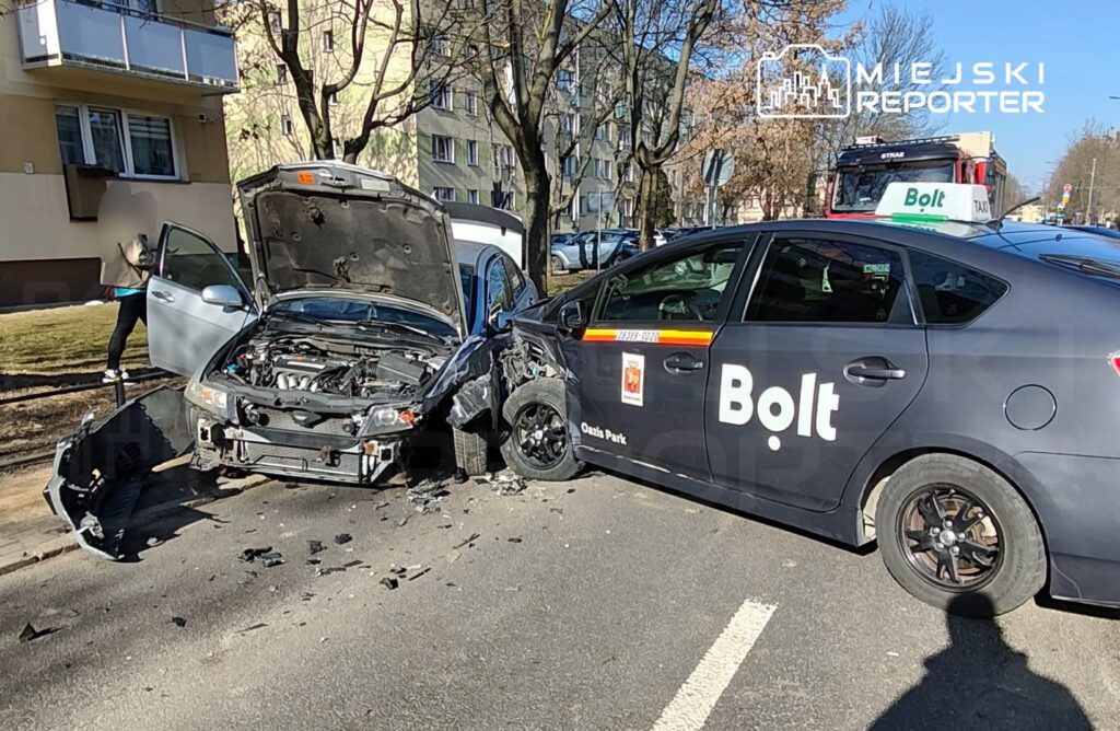 Zniszczony samochód osobowy z otwartą maską stoi obok uszkodzonego pojazdu z logo Bolt na ulicy w miejskim otoczeniu.