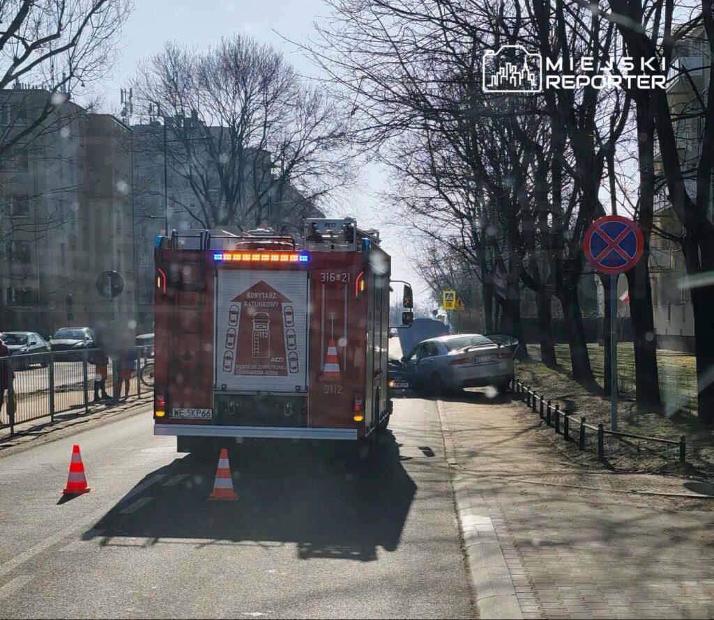 Straż pożarna z włączonymi sygnałami świetlnymi zatrzymana obok uszkodzonego samochodu na chodniku.