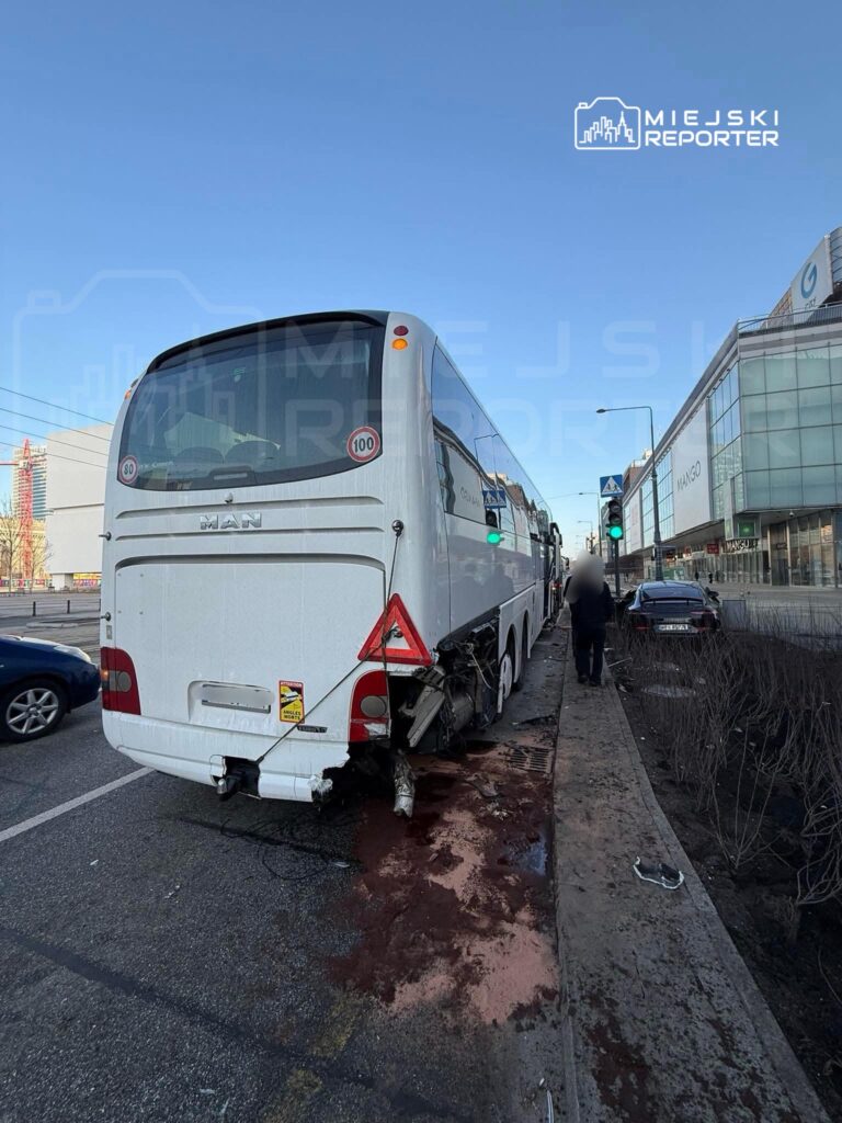 Uszkodzony autobus stojący na poboczu, z widocznymi śladami uszkodzeń z tyłu, obok ruchliwej ulicy.