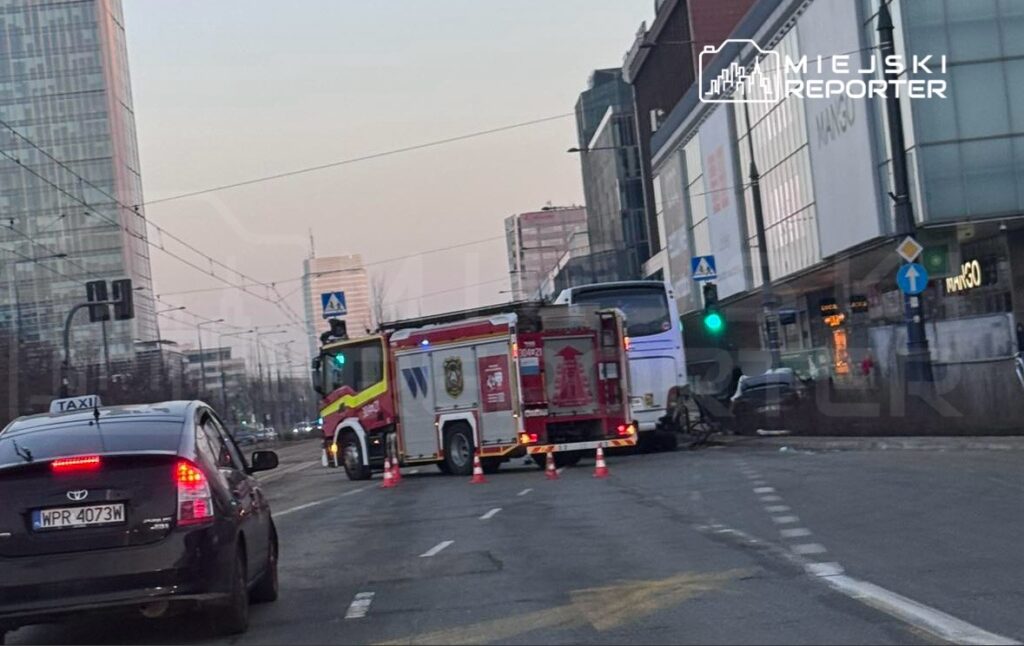 Wozidło strażackie z włączonymi sygnałami stoi obok zderzonego autobusu na ruchliwej ulicy w centrum miasta.
