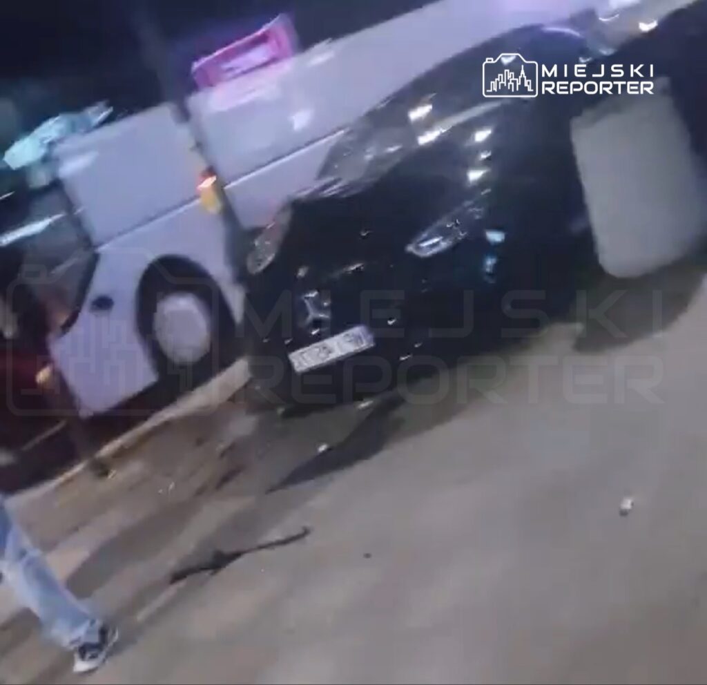 Czarny samochód osobowy stojący na jezdni obok białego autobusu, w tle widoczna nocna sceneria uliczna.