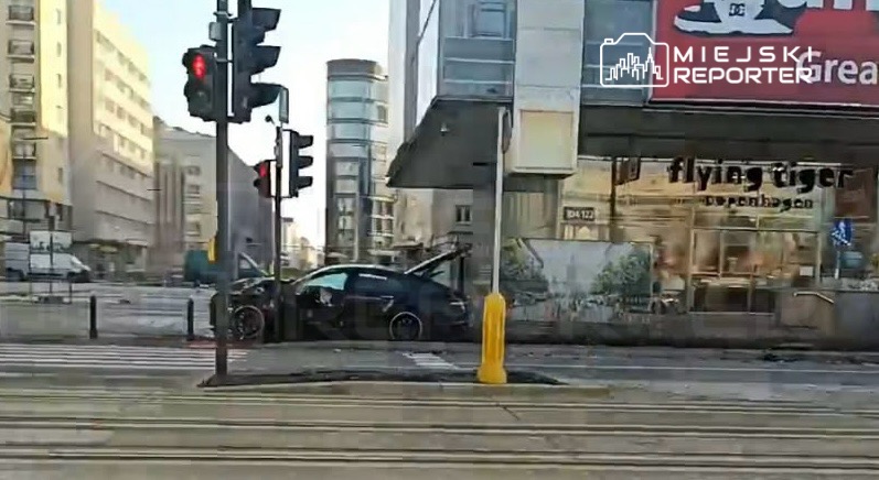 Czarny samochód z otwartymi drzwiami stoi na ulicy przed nowoczesnym budynkiem, obok sygnalizacji świetlnej.