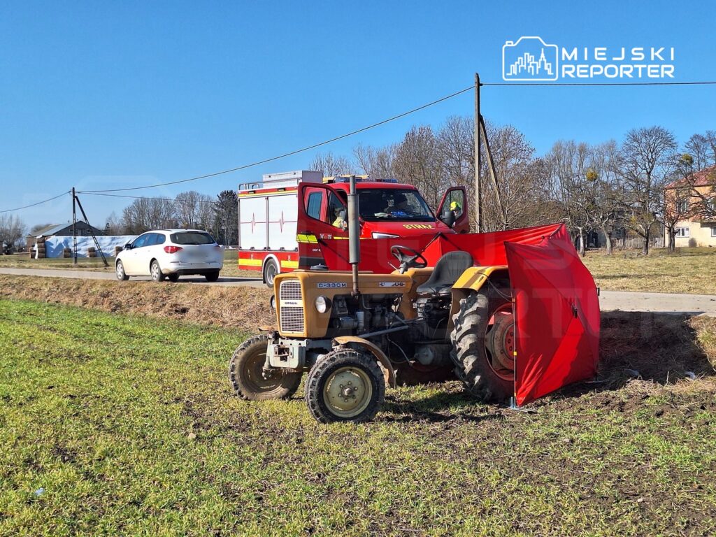 Traktor z czerwoną osłoną stoi na polu obok wozu strażackiego, a w tle widać wiejską drogę i domy.