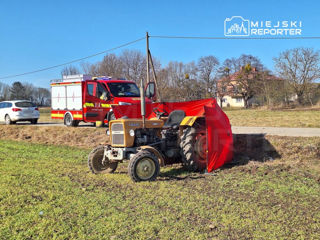 Traktor z czerwoną osłoną stoi na polu obok drogi, a w tle widoczny jest wóz strażacki oraz dom.