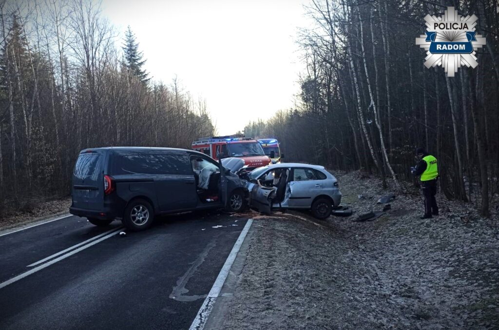 Zderzenie dwóch samochodów na drodze, obok stojący radiowóz i wozem strażackim, w otoczeniu lasu.