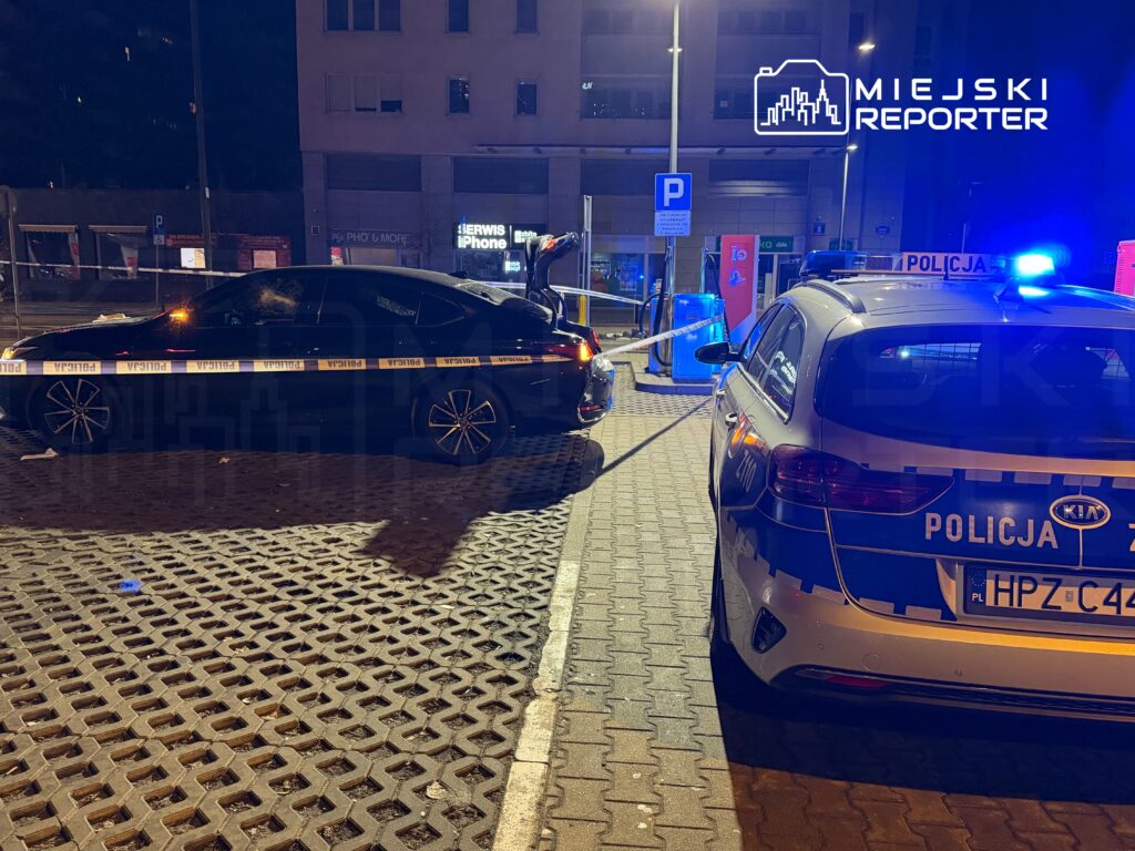 Czarny samochód osobowy z policyjnym taśmą zabezpieczającą oraz radiowóz z włączonymi sygnałami świetlnymi na ulicy nocą.