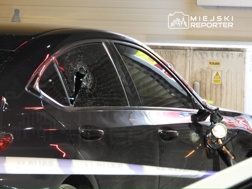 Czarny samochód osobowy z wybitym tylnym oknem stoi na parkingu, obok widoczna taśma policyjna.