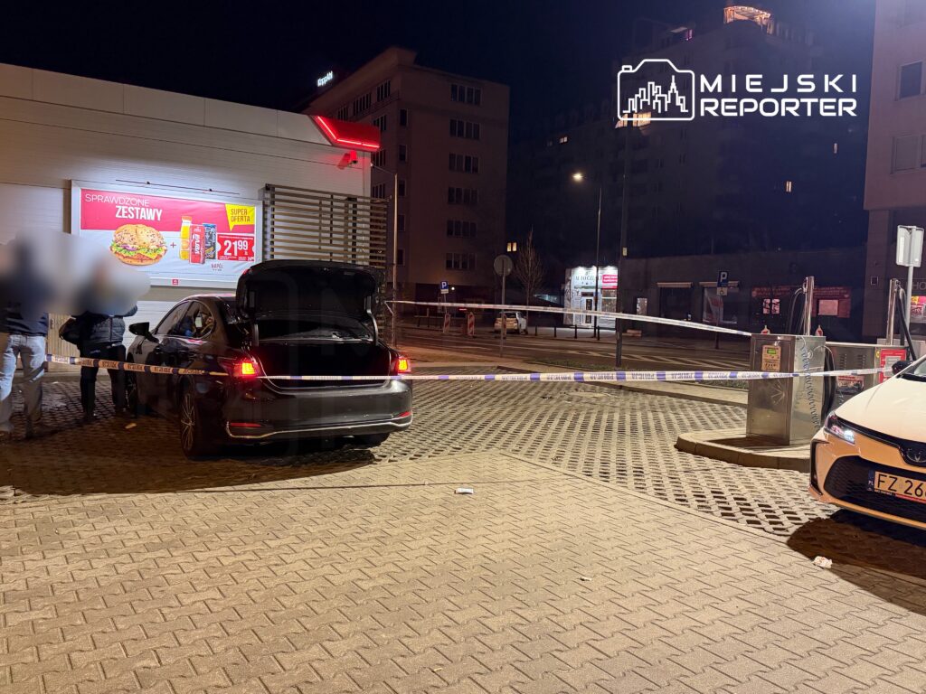 Czarna limuzyna z otwartą klapą bagażnika stoi na parkingu, otoczona taśmą policyjną. W tle widoczny jest jasny budynek z reklamą.