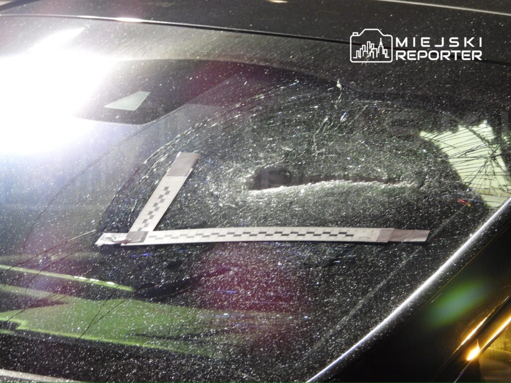 Pęknięta szyba samochodu z oznaczeniem i linijką pomiarową na tle nocnego oświetlenia.