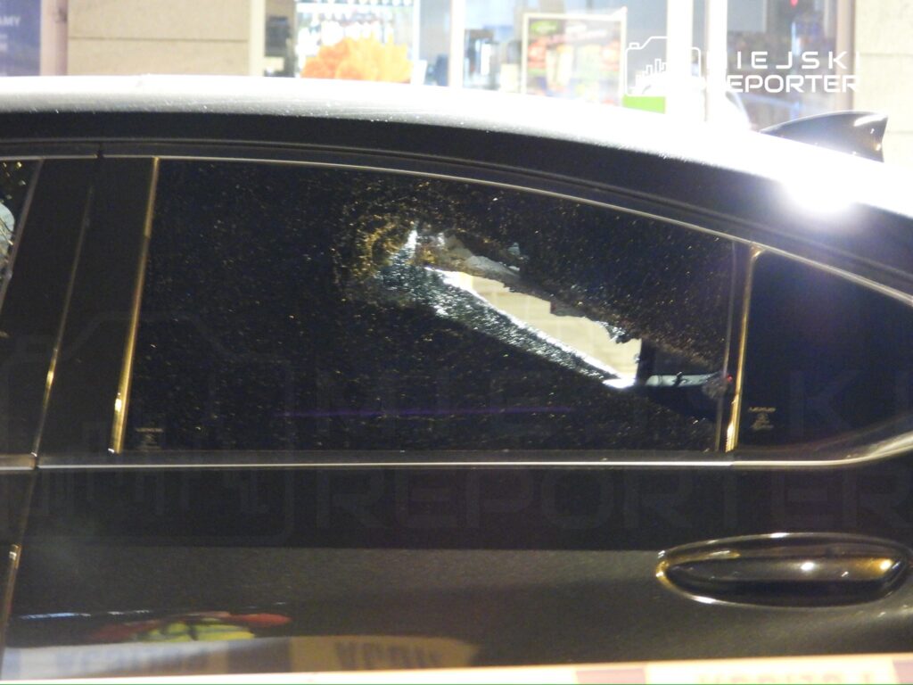 Uszkodzona szyba boczna czarnego samochodu zaparkowanego w miejskim otoczeniu. W tle widoczne oświetlone witryny sklepowe.