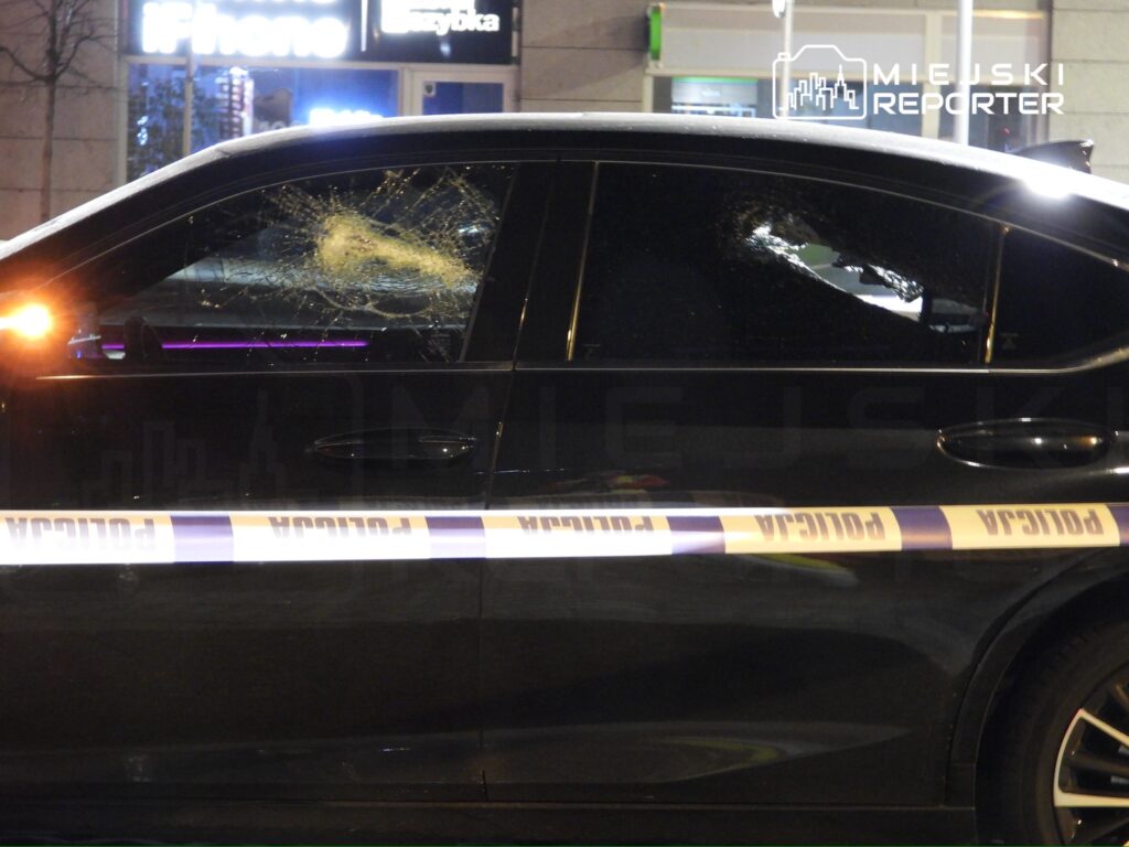 Zniszczona szyba w drzwiach czarnego samochodu osobowego, zabezpieczona taśmą policyjną na tle oświetlonych witryn sklepowych.