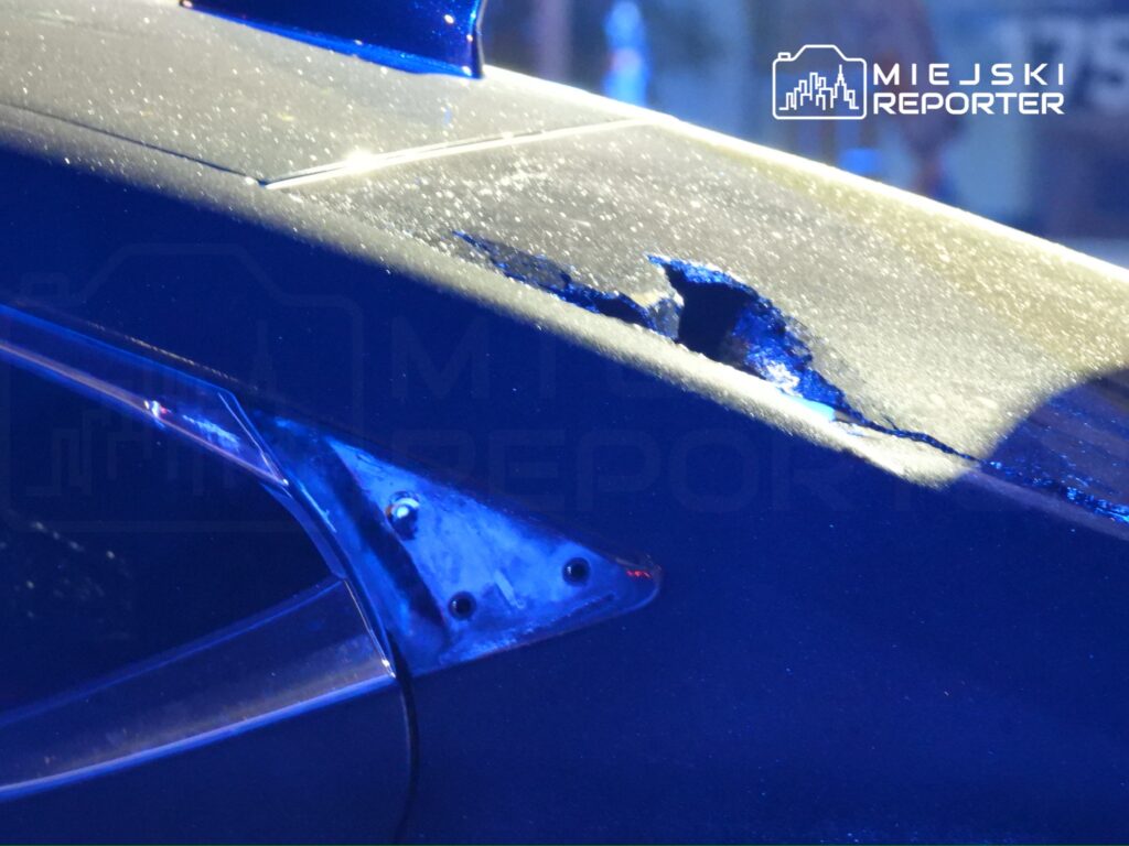 Uszkodzona karoseria samochodu z widoczną rysą i odłamanym lakierem, oświetlona niebieskim światłem.