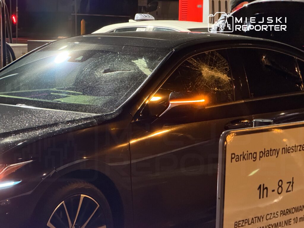 Uszkodzony czarny samochód z pękniętą szybą, zaparkowany na oświetlonym parkingu z tablicą informacyjną o opłatach.