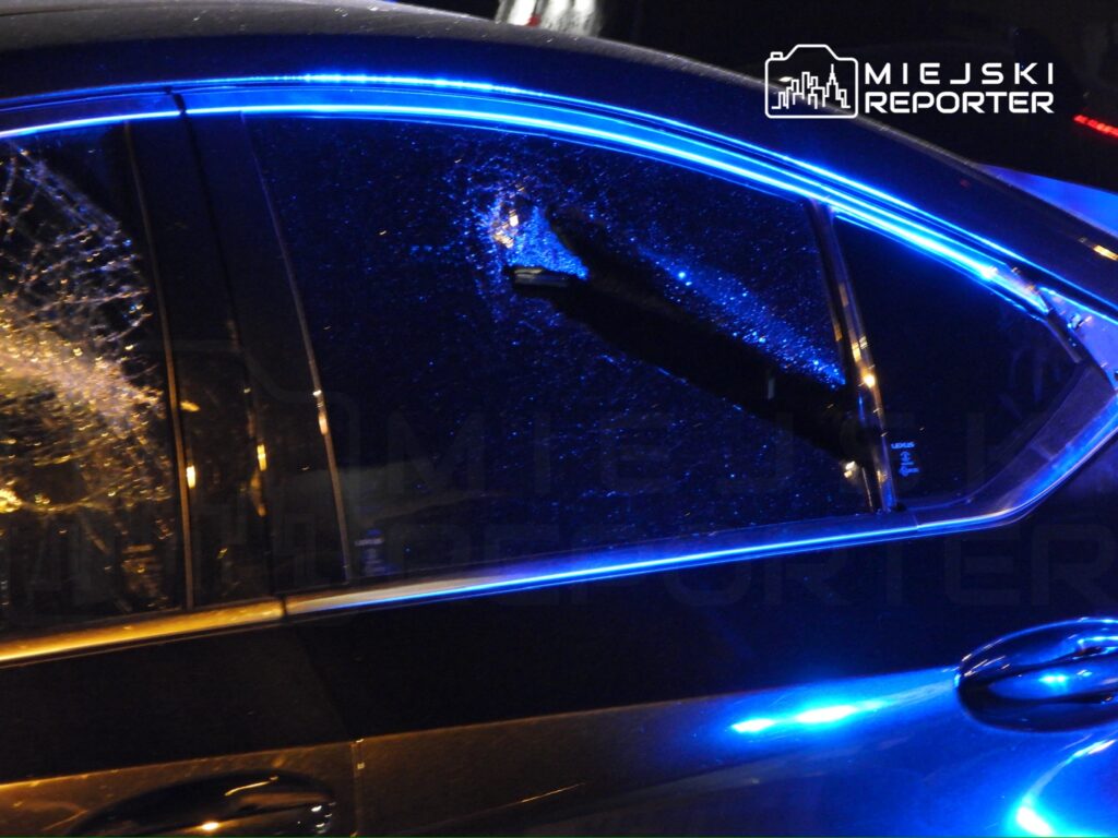 Zniszczona szyba samochodu z pęknięciami, oświetlona niebieskim światłem w nocnej scenerii.